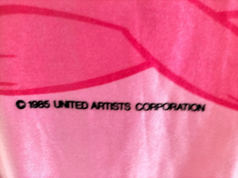 Pode incluir: Tecido rosa com um design rosa mais escuro. O texto "&copy; 1985 UNITED ARTISTS CORPORATION" est&aacute; impresso em preto na parte inferior da imagem. O tecido tem uma textura suave.