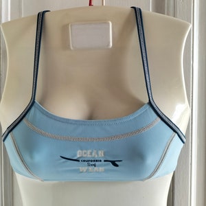 Op de afbeelding: Lichtblauwe sportbeha met smalle marineblauwe bandjes en witte stiksels. De beha heeft de tekst "OCEAN CALIFORNIA SURF WEAR" met een surfplankafbeelding. De beha wordt getoond op een mannequin.