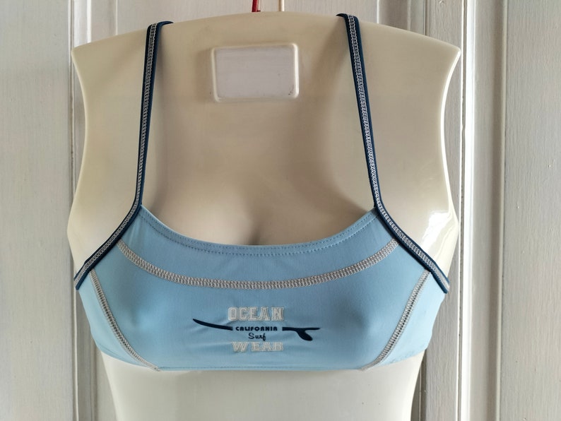 Op de afbeelding: Lichtblauwe sportbeha met smalle marineblauwe bandjes en witte stiksels. De beha heeft de tekst "OCEAN CALIFORNIA SURF WEAR" met een surfplankafbeelding. De beha wordt getoond op een mannequin.
