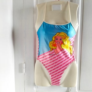 Puede incluir: Un traje de ba&ntilde;o rosa y azul con un patr&oacute;n de rayas y un gr&aacute;fico de una mu&ntilde;eca rubia con ojos azules.