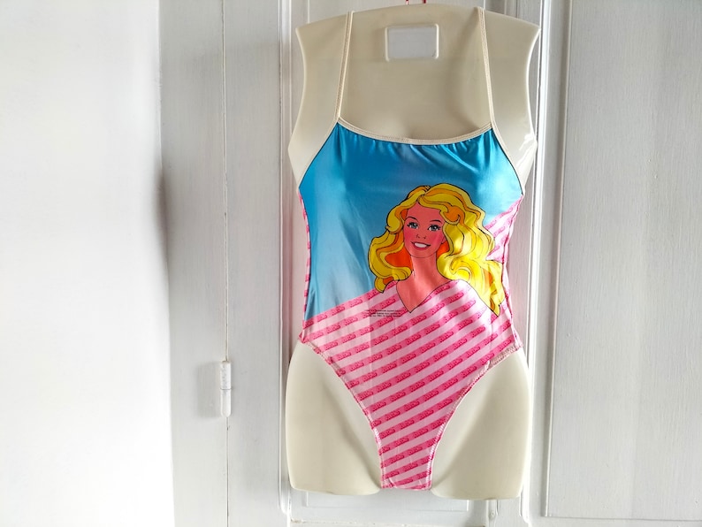 Puede incluir: Un traje de ba&ntilde;o rosa y azul con un patr&oacute;n de rayas y un gr&aacute;fico de una mu&ntilde;eca rubia con ojos azules.