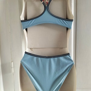 Op de afbeelding: Lichtblauwe bikiniset met zwart-witte bies. De top heeft een racerback-ontwerp en de high-waisted broekjes bieden volledige bedekking. De badkleding wordt gepresenteerd op een mannequin tegen een witte achtergrond.