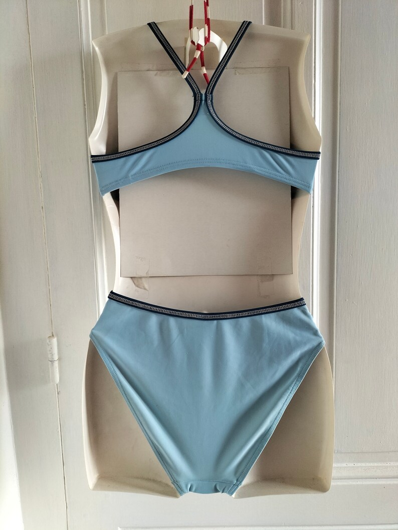 Op de afbeelding: Lichtblauwe bikiniset met zwart-witte bies. De top heeft een racerback-ontwerp en de high-waisted broekjes bieden volledige bedekking. De badkleding wordt gepresenteerd op een mannequin tegen een witte achtergrond.