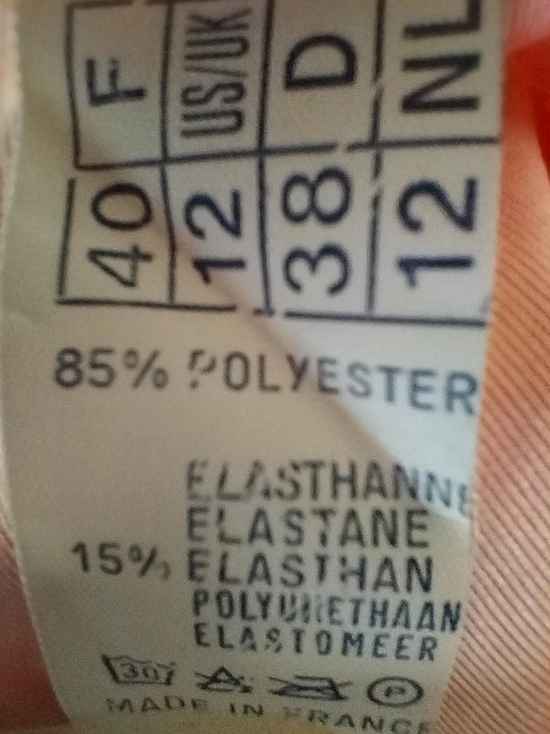 Pode incluir: Close-up de uma etiqueta de roupa com informa&ccedil;&otilde;es de tamanho e composi&ccedil;&atilde;o do tecido. A etiqueta mostra tamanhos para Fran&ccedil;a (40), US/UK (12), Alemanha (38) e Pa&iacute;ses Baixos (12). Lista 85% poli&eacute;ster e 15% elastano. A etiqueta &eacute; marcada "Made in France."