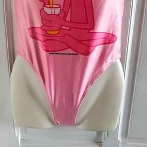 Pode incluir: Fato de banho rosa de uma pe&ccedil;a com um gr&aacute;fico da Pantera Cor-de-Rosa segurando uma bebida. O fato de banho &eacute; exibido em um manequim. O texto "&copy; 1968 UNITED ARTISTS CORPORATION" est&aacute; impresso no fato de banho.