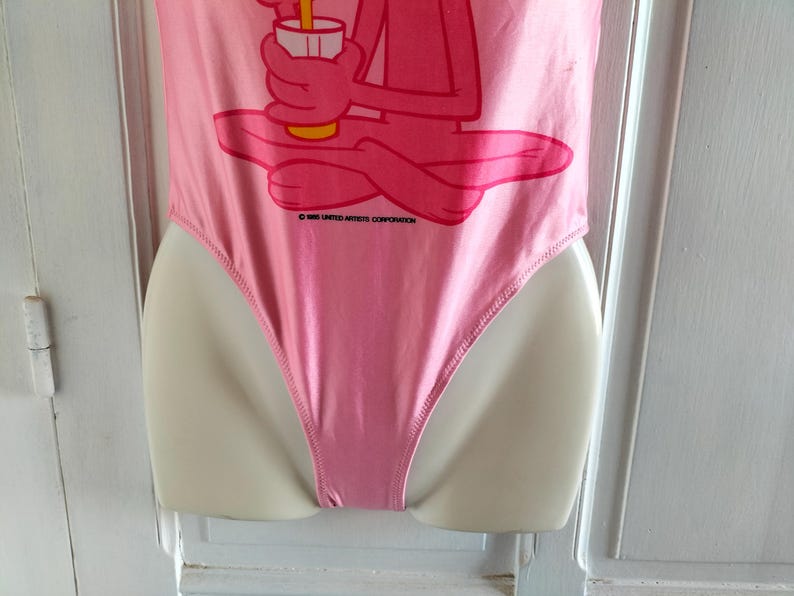 Pode incluir: Fato de banho rosa de uma pe&ccedil;a com um gr&aacute;fico da Pantera Cor-de-Rosa segurando uma bebida. O fato de banho &eacute; exibido em um manequim. O texto "&copy; 1968 UNITED ARTISTS CORPORATION" est&aacute; impresso no fato de banho.