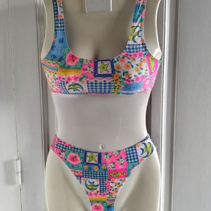 Puede incluir: Un traje de ba&ntilde;o de dos piezas con un dise&ntilde;o de patchwork colorido. La parte superior es de estilo cuello redondo, y la parte inferior es de corte alto. El patr&oacute;n incluye motivos florales, delfines y estrellas en rosa, azul y amarillo. El traje de ba&ntilde;o se muestra en un maniqu&iacute;.