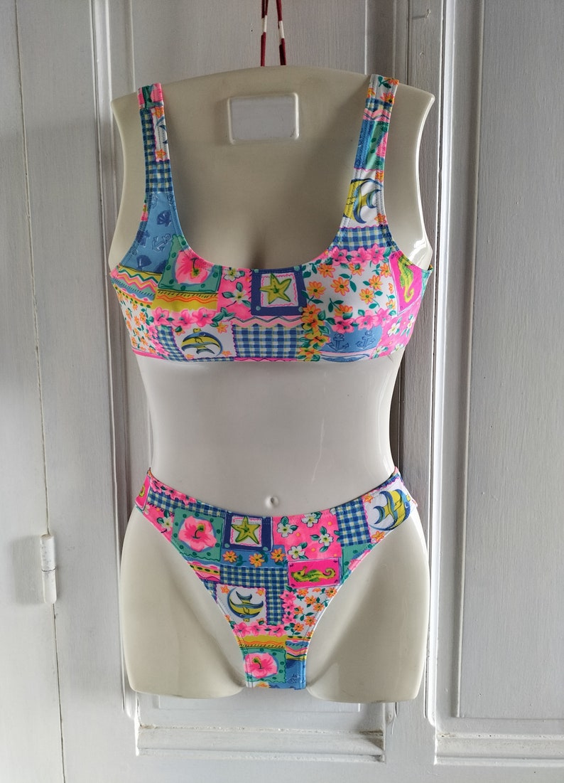 Puede incluir: Un traje de ba&ntilde;o de dos piezas con un dise&ntilde;o de patchwork colorido. La parte superior es de estilo cuello redondo, y la parte inferior es de corte alto. El patr&oacute;n incluye motivos florales, delfines y estrellas en rosa, azul y amarillo. El traje de ba&ntilde;o se muestra en un maniqu&iacute;.