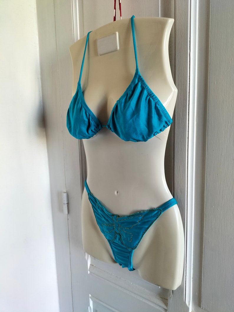 May include: A turquoise bikini with a butterfly design on the bottom. The bikini is displayed on a white mannequin.