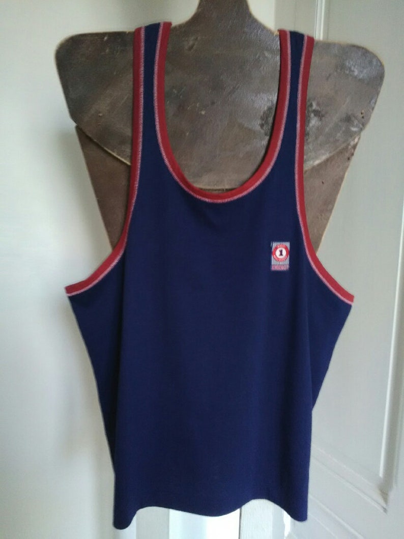 May include: A navy blue sleeveless shirt with red and white trim around the neck and armholes. The shirt has a small logo on the front with a red circle and the number "1" inside.