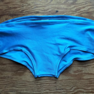 Könnte beinhalten: Eine hellblaue Badehose. Die Hose besteht aus einem glatten Stoff und hat einen hoch taillierten Schnitt. Die Nähte sind sichtbar und mit passendem Faden genäht. Die Bademode wird auf einer Holzoberfläche präsentiert.