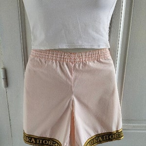 Puede incluir: Pantalones cortos deportivos de color rosa con cintura el&aacute;stica dorada y ribete dorado con el texto "КА  ПОБЕЗ  КА  ПОБЕЗ" en la parte inferior.