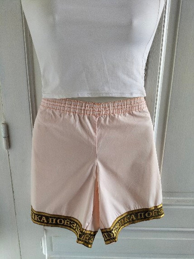 Puede incluir: Pantalones cortos deportivos de color rosa con cintura el&aacute;stica dorada y ribete dorado con el texto "КА  ПОБЕЗ  КА  ПОБЕЗ" en la parte inferior.