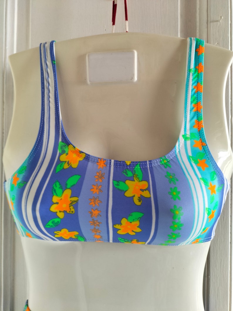 Op de afbeelding: Een blauw en wit gestreept badpak bovenstuk met een bloemenpatroon. De bloemen zijn oranje, geel en groen.