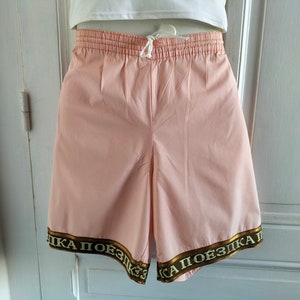 Puede incluir: Pantalones cortos de color rosa con un ribete dorado y blanco que dice "КА ПОЕЗДКА П".