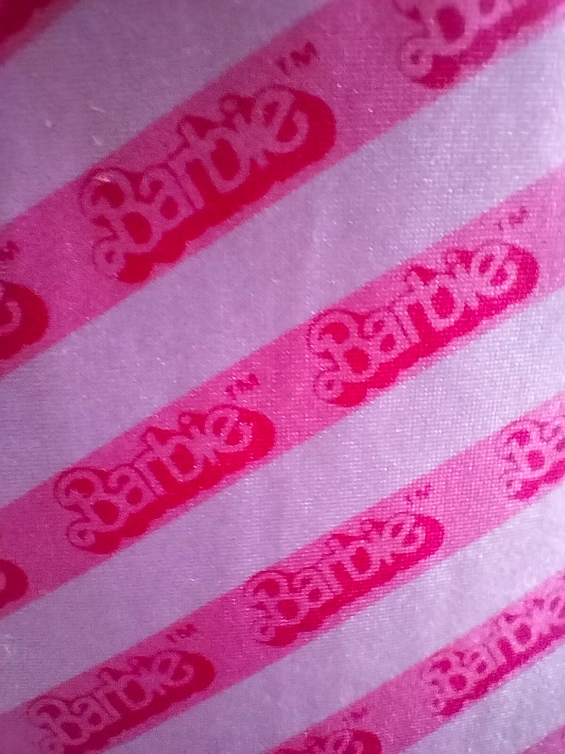 Puede incluir: Tela de rayas rosa y blanca con la palabra "Barbie" repetida en letras rosas.