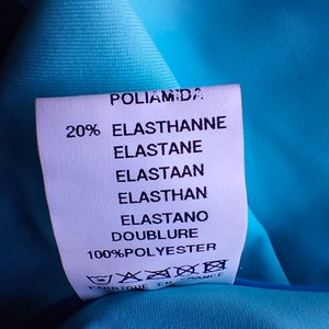 May include: A white fabric label with black text detailing the fabric composition of a garment. The label reads "Poliamida 20% Elasthanne Elastane Elastaan Elasthan Elastano Doublure 100%Polyester" and includes a care label with symbols for washing, ironing, and dry cleaning. The label is made in France.