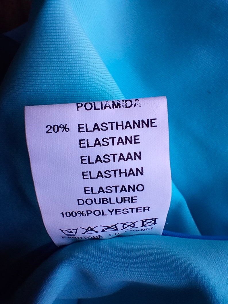 May include: A white fabric label with black text detailing the fabric composition of a garment. The label reads "Poliamida 20% Elasthanne Elastane Elastaan Elasthan Elastano Doublure 100%Polyester" and includes a care label with symbols for washing, ironing, and dry cleaning. The label is made in France.