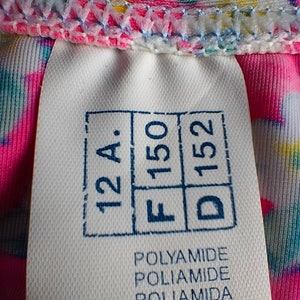 Puede incluir: Primer plano de una etiqueta blanca con texto y n&uacute;meros estampados en azul, incluyendo "12 A", "F 150" y "D 152". La etiqueta tambi&eacute;n enumera "POLYAMIDE" en varios idiomas y "80 %". La etiqueta est&aacute; adherida a una tela colorida.