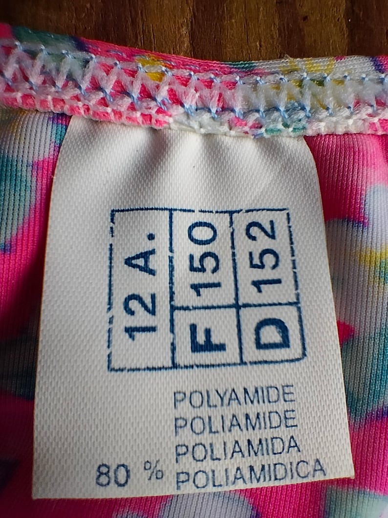 Puede incluir: Primer plano de una etiqueta blanca con texto y n&uacute;meros estampados en azul, incluyendo "12 A", "F 150" y "D 152". La etiqueta tambi&eacute;n enumera "POLYAMIDE" en varios idiomas y "80 %". La etiqueta est&aacute; adherida a una tela colorida.