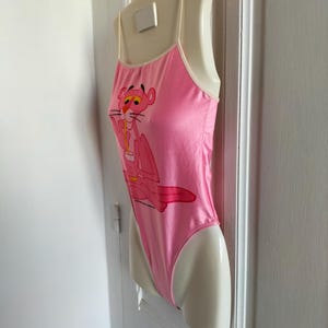 Pode incluir: Body rosa com al&ccedil;as finas e acabamento branco. O body apresenta um desenho da Pantera Cor-de-Rosa bebendo de uma x&iacute;cara. A pe&ccedil;a de roupa &eacute; exibida em um manequim.
