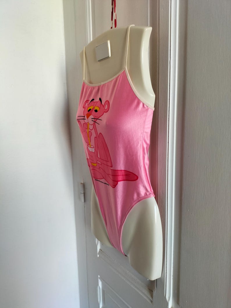 Pode incluir: Body rosa com al&ccedil;as finas e acabamento branco. O body apresenta um desenho da Pantera Cor-de-Rosa bebendo de uma x&iacute;cara. A pe&ccedil;a de roupa &eacute; exibida em um manequim.