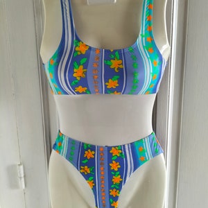 Op de afbeelding: Een blauwe en witte gestreepte bikini met oranje bloemen. De bikini top heeft een ronde hals en dunne bandjes. De bikini broek is een hoog uitgesneden model.