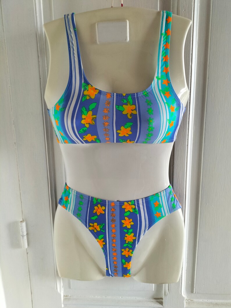 Op de afbeelding: Een blauwe en witte gestreepte bikini met oranje bloemen. De bikini top heeft een ronde hals en dunne bandjes. De bikini broek is een hoog uitgesneden model.