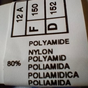 Op de afbeelding: Een wit kledinglabel met zwarte tekst. Het label toont de maten 12A, F 150 en D 152. Het label vermeldt ook "80% Polyamide, Nylon, Polyamid, Poliamida, Poliam&igrave;dica, Poliamida."
