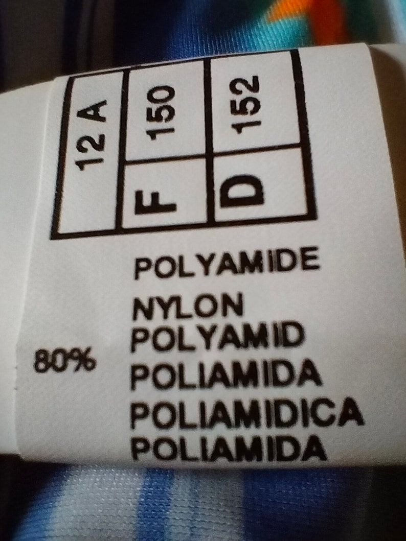 Op de afbeelding: Een wit kledinglabel met zwarte tekst. Het label toont de maten 12A, F 150 en D 152. Het label vermeldt ook "80% Polyamide, Nylon, Polyamid, Poliamida, Poliam&igrave;dica, Poliamida."