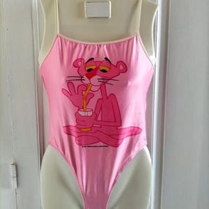 Pode incluir: Fato de banho rosa de uma pe&ccedil;a com um desenho da Pantera Cor-de-Rosa a beber de um copo. O fato de banho tem al&ccedil;as finas e um decote redondo. A Pantera Cor-de-Rosa est&aacute; sentada. O fato de banho &eacute; exibido num manequim.