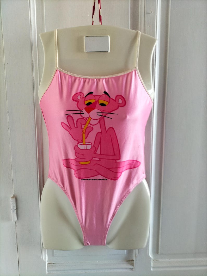 Pode incluir: Fato de banho rosa de uma pe&ccedil;a com um desenho da Pantera Cor-de-Rosa a beber de um copo. O fato de banho tem al&ccedil;as finas e um decote redondo. A Pantera Cor-de-Rosa est&aacute; sentada. O fato de banho &eacute; exibido num manequim.