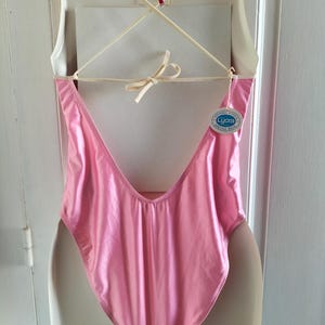 Pode incluir: Um fato de banho cor-de-rosa brilhante, de uma pe&ccedil;a, com as costas baixas e pernas cavadas. O fato de banho est&aacute; pendurado num cabide branco. Uma etiqueta com a palavra "Lycra" est&aacute; presa.