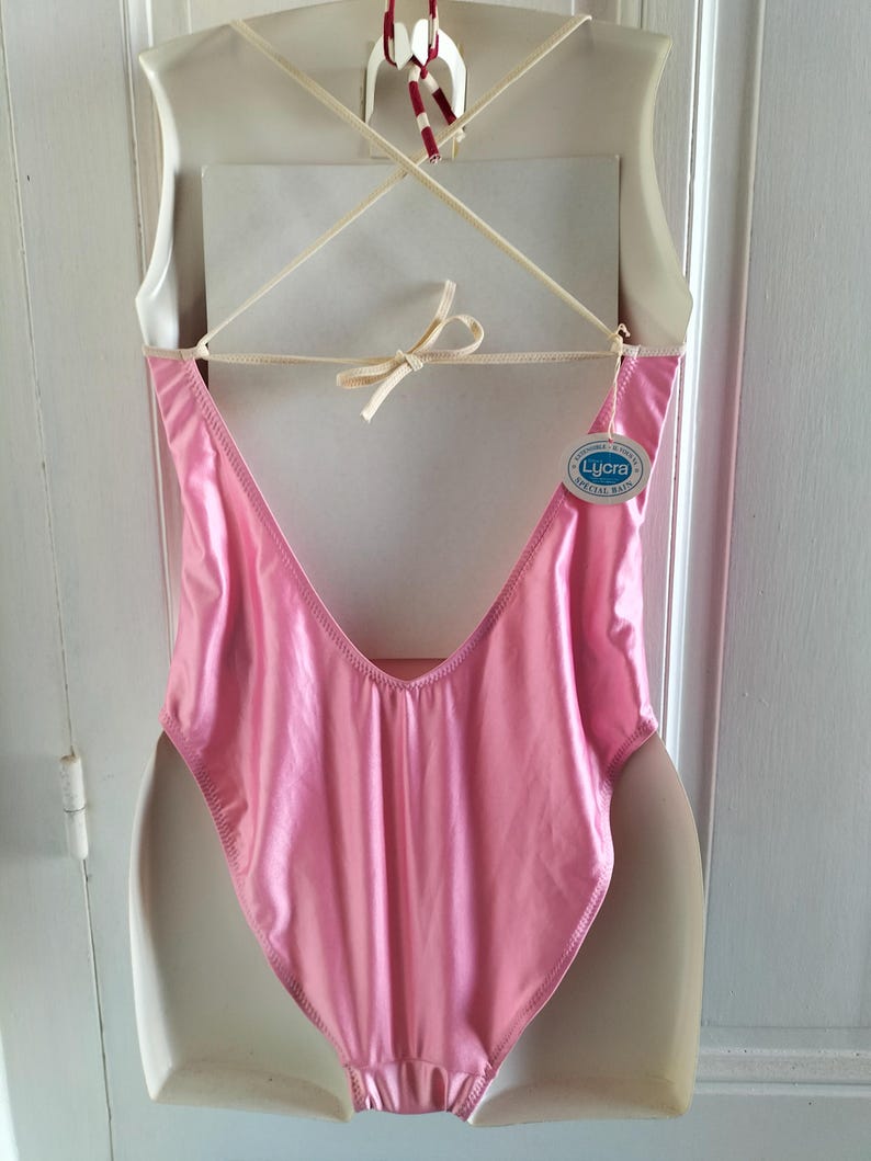 Pode incluir: Um fato de banho cor-de-rosa brilhante, de uma pe&ccedil;a, com as costas baixas e pernas cavadas. O fato de banho est&aacute; pendurado num cabide branco. Uma etiqueta com a palavra "Lycra" est&aacute; presa.