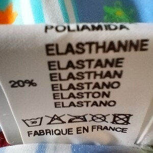 Op de afbeelding: Een wit stoffen label met zwarte tekst die "Poliamida Elastthanne Elastane Elasthan Elastano Elaston Elastano" en "20%" luidt. Het label bevat ook een wasinstructie symbool en de tekst "Fabrique en France".