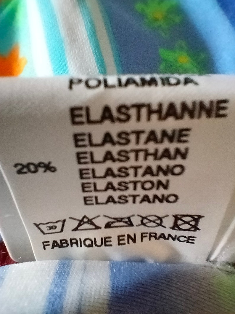 Op de afbeelding: Een wit stoffen label met zwarte tekst die "Poliamida Elastthanne Elastane Elasthan Elastano Elaston Elastano" en "20%" luidt. Het label bevat ook een wasinstructie symbool en de tekst "Fabrique en France".
