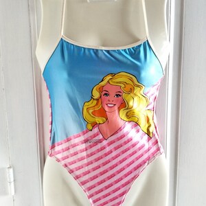 Puede incluir: Un traje de ba&ntilde;o rosa y azul con un gr&aacute;fico de una mu&ntilde;eca Barbie. El traje de ba&ntilde;o tiene un ribete blanco y un dise&ntilde;o de tirantes finos. La mu&ntilde;eca Barbie lleva una camisa a rayas rosa y blanca.