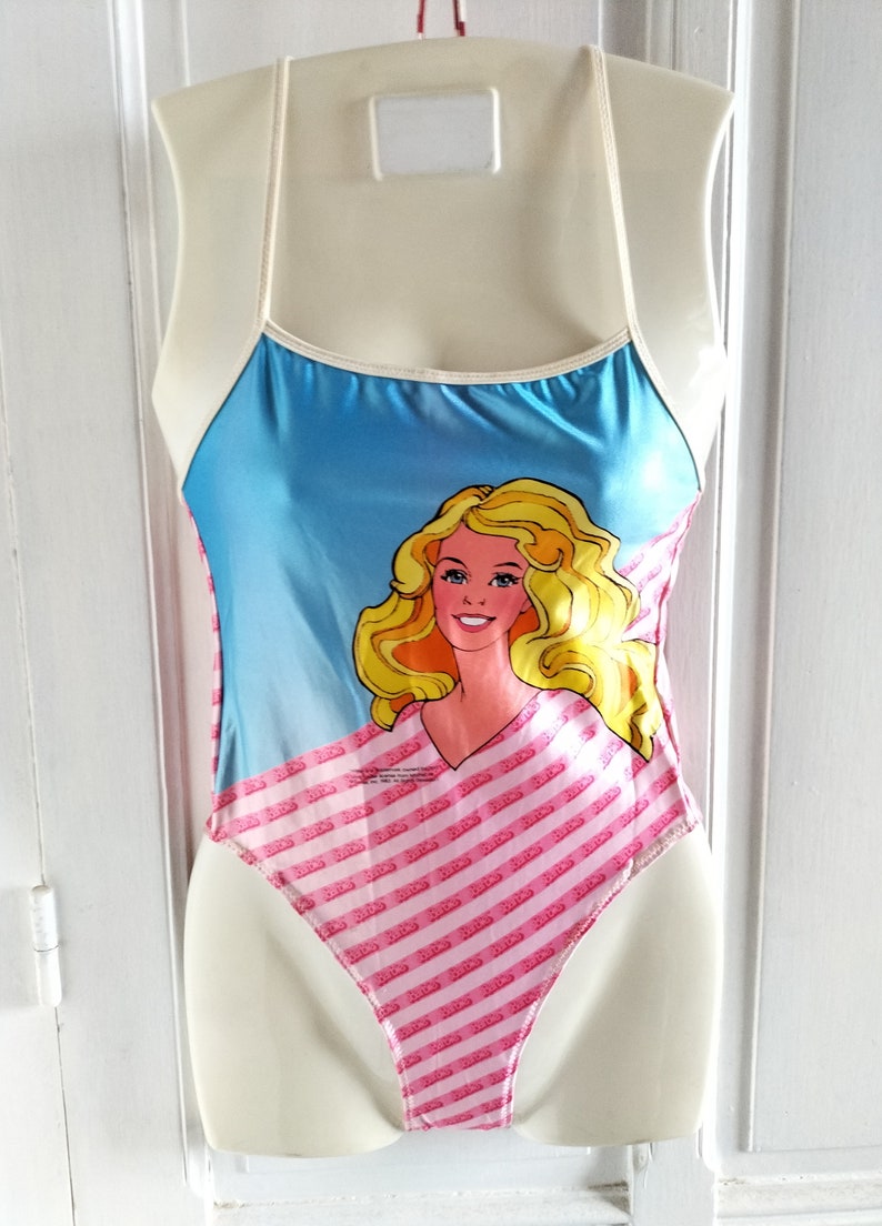Puede incluir: Un traje de ba&ntilde;o rosa y azul con un gr&aacute;fico de una mu&ntilde;eca Barbie. El traje de ba&ntilde;o tiene un ribete blanco y un dise&ntilde;o de tirantes finos. La mu&ntilde;eca Barbie lleva una camisa a rayas rosa y blanca.