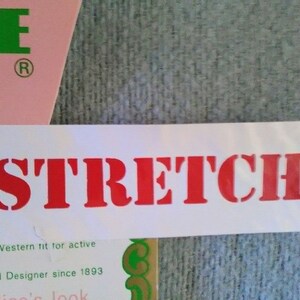 May include: White label with the word "STRETCH" printed in red letters. The label is attached to a pink and green fabric with the text "CE" and "Makers and Designer since 1893".