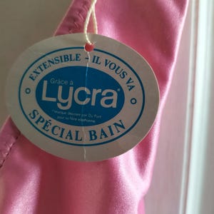 Pode incluir: Um close-up de uma pe&ccedil;a de roupa rosa com uma etiqueta redonda anexada. A etiqueta tem o texto "EXTENSIBLE - IL VOUS VA" e "SP&Eacute;CIAL BAIN" com a palavra "Lycra" no centro. A pe&ccedil;a de roupa &eacute; feita de um material brilhante e liso.