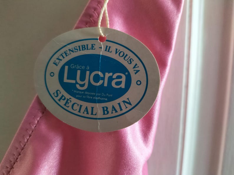 Pode incluir: Um close-up de uma pe&ccedil;a de roupa rosa com uma etiqueta redonda anexada. A etiqueta tem o texto "EXTENSIBLE - IL VOUS VA" e "SP&Eacute;CIAL BAIN" com a palavra "Lycra" no centro. A pe&ccedil;a de roupa &eacute; feita de um material brilhante e liso.