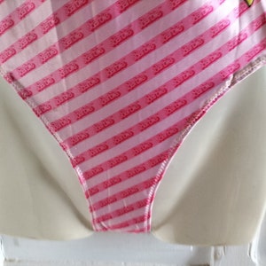 Puede incluir: Traje de ba&ntilde;o rosa y blanco con un patr&oacute;n repetido de la palabra "Barbie" en rosa. El traje de ba&ntilde;o tiene una pierna de corte alto y una espalda baja.