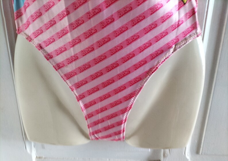 Puede incluir: Traje de ba&ntilde;o rosa y blanco con un patr&oacute;n repetido de la palabra "Barbie" en rosa. El traje de ba&ntilde;o tiene una pierna de corte alto y una espalda baja.