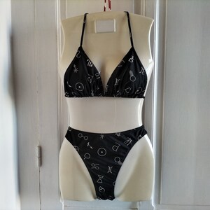 May include: Black bikini with a white astrological symbol print. The top is a triangle style with thin straps. The bottom is a high-cut style.