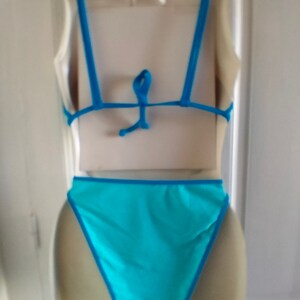 May include: A turquoise blue swimsuit with thin blue straps and a tie back. The swimsuit has a high-cut leg and a low back.