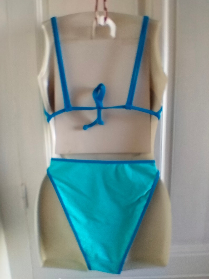 May include: A turquoise blue swimsuit with thin blue straps and a tie back. The swimsuit has a high-cut leg and a low back.