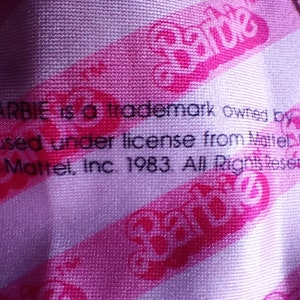 Puede incluir: Una tela de rayas rosa y blancas con el texto "Barbie is a trademark owned by and used under license from Mattel, Inc. 1983. All Rights Reserved." y el logotipo de Barbie en rosa.