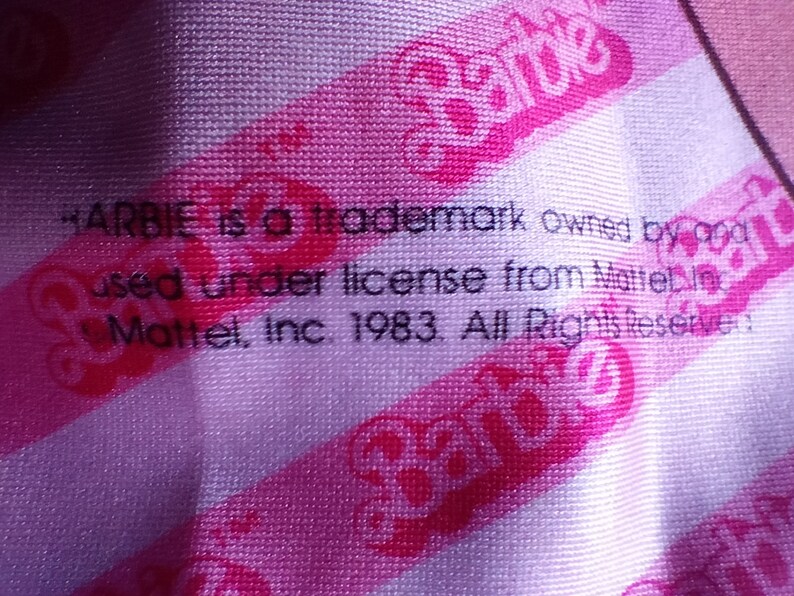 Puede incluir: Una tela de rayas rosa y blancas con el texto "Barbie is a trademark owned by and used under license from Mattel, Inc. 1983. All Rights Reserved." y el logotipo de Barbie en rosa.