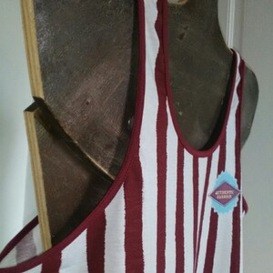 May include: A white and maroon striped tank top with a red trim around the neckline. The tank top is hanging on a wooden hanger. The tag on the tank top says "Authentic Fashion".