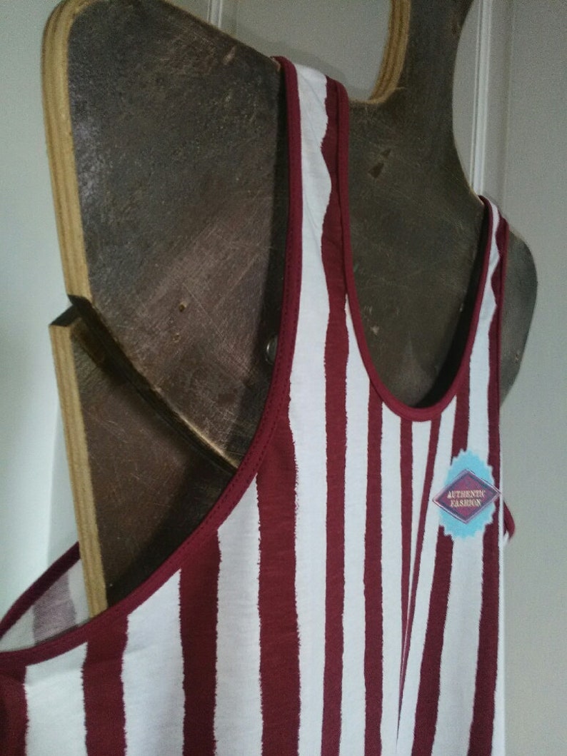 May include: A white and maroon striped tank top with a red trim around the neckline. The tank top is hanging on a wooden hanger. The tag on the tank top says "Authentic Fashion".
